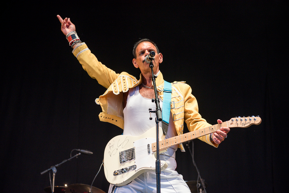 Freddie Mercury on a stage in front of the microphone, wearing a golden jacket with a guiter in his hand 