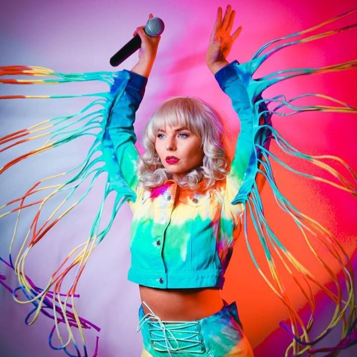 Blonde woman is standing with her arms up, rainbow coloured strings falling from her arms